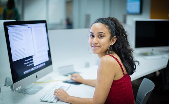 Student sitting at a large computer monitor, looking back over her shoulder and smiling at camera