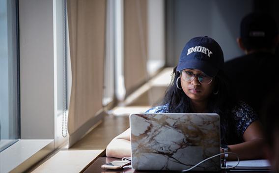 Student in Emory cap working on a laptop in the Woodruff Library