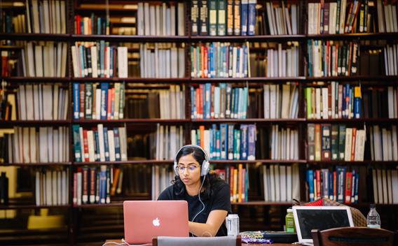 Student at her laptop with headphones, book shelves in the background